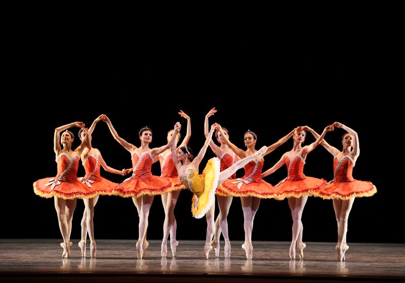 Soloist Sarah Lane in a bright yellow tutu strikes a high arabesque while the corps de ballet in orange tutus stand en pointe in the background